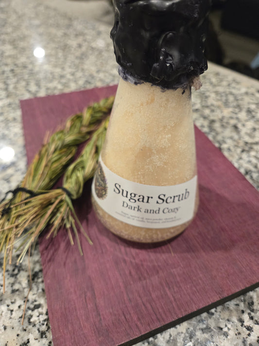 Bottle of sugar scrub labeled 'Dark and Cozy' on a purple towel with a black bar of soap and dried herbs.